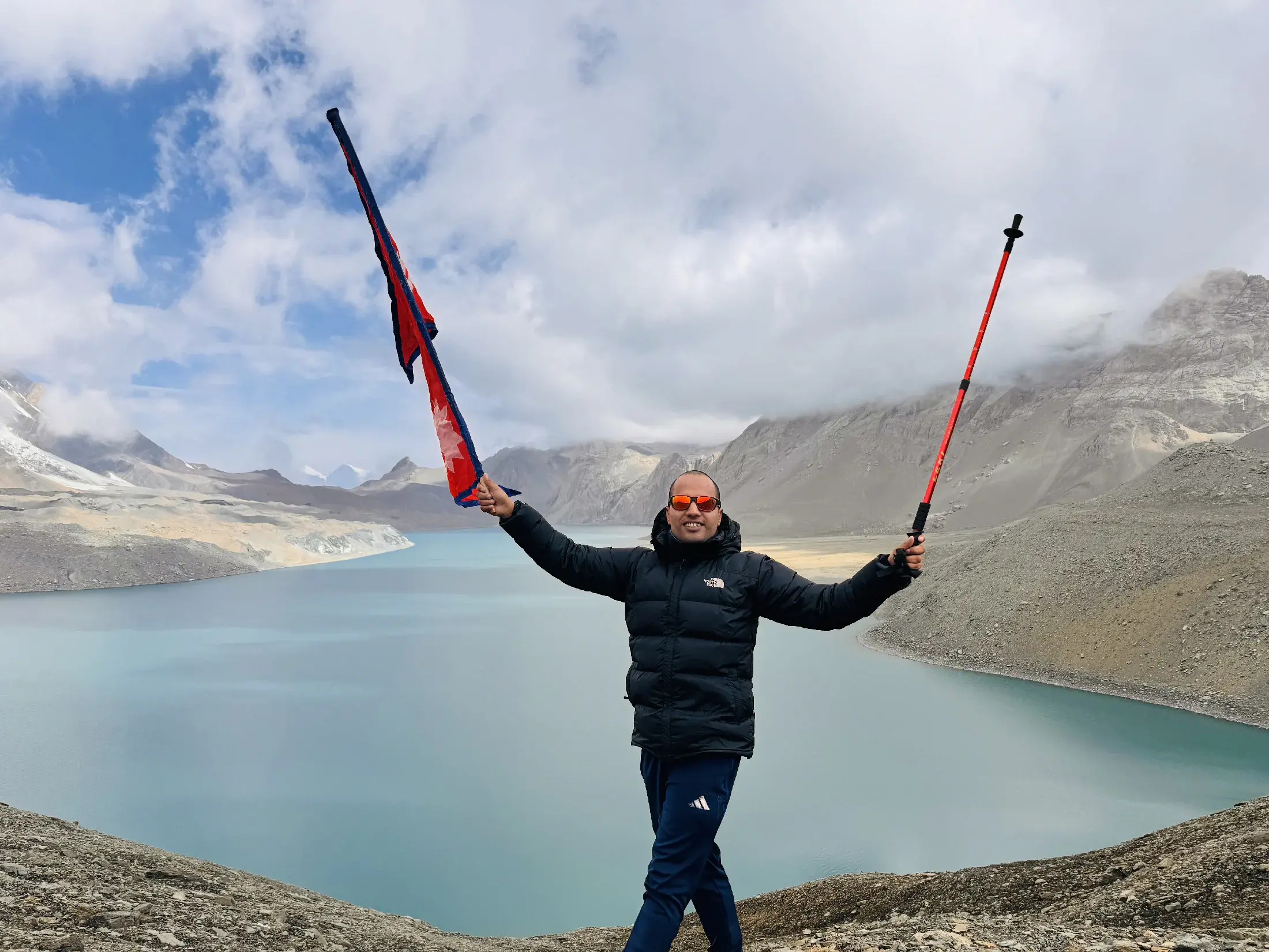 Khagendra holding Nepal's flag and a trekking pole above a turquoise Himalayan lake